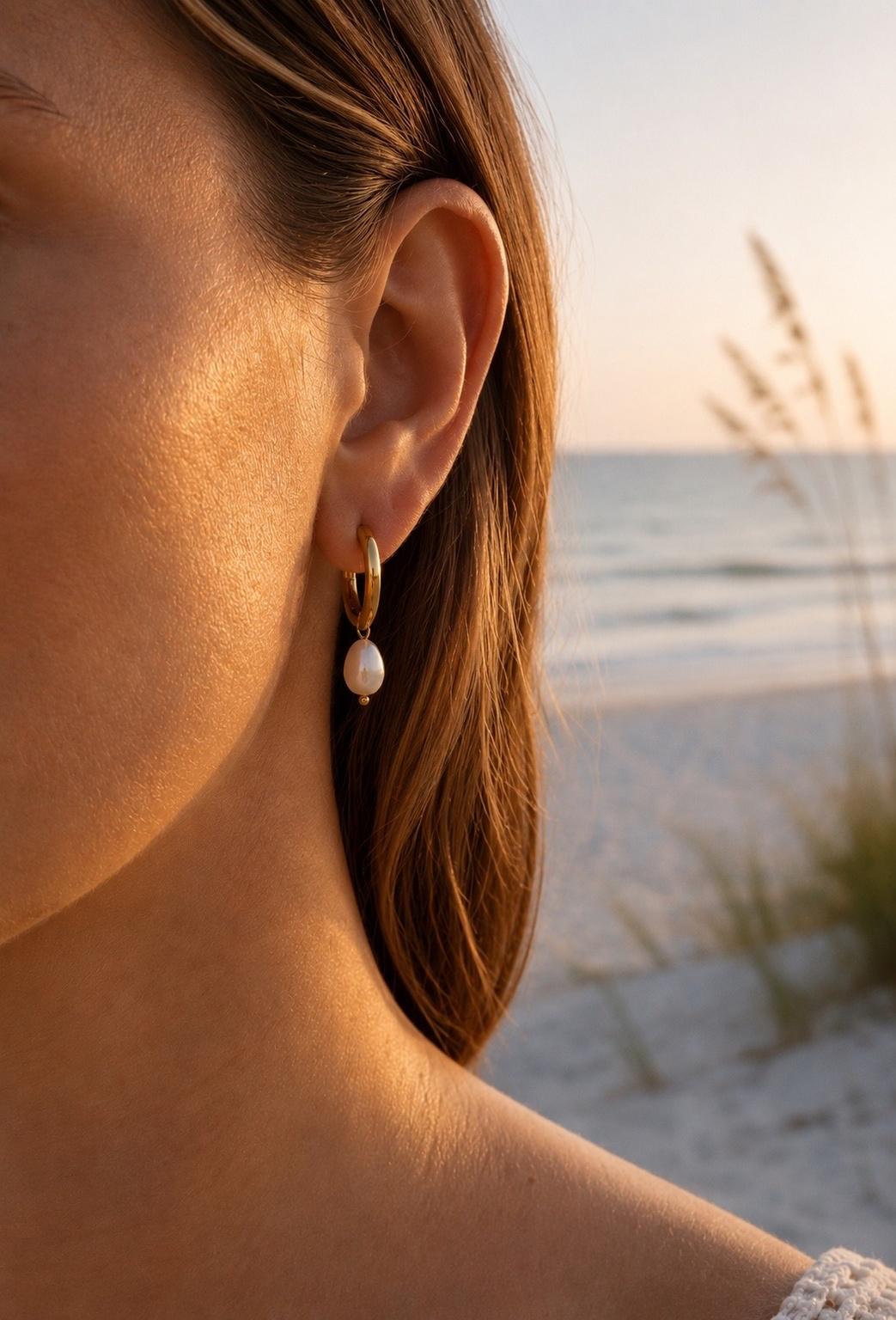 Goldene Creole mit einer echten Barockperle als Anhänger von hejschmuck, getragen am Ohr einer Frau am Strand bei Sonnenuntergang.