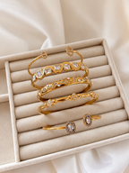 Ariella Armreif in Gold mit Zirkonia, gestylt mit weiteren goldenen Armreifen in einer cremefarbenen Samtschale