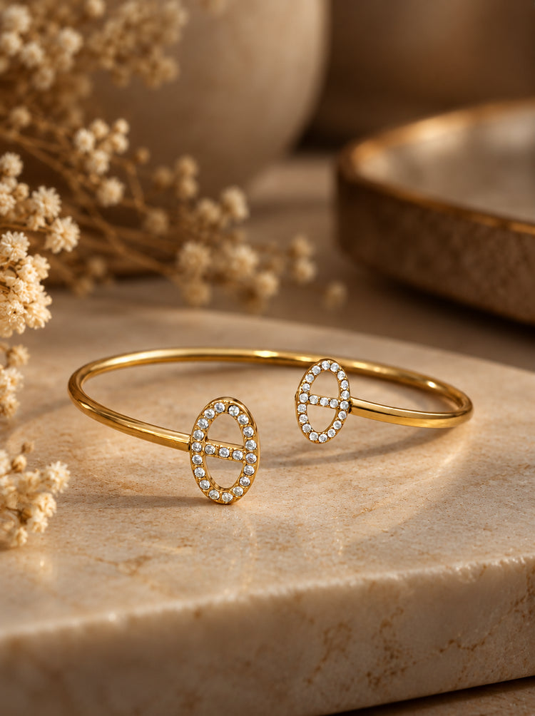 Athena Armreif in Gold mit ovalen Zirkonia-Enden auf heller Steinplatte neben Trockenblumen