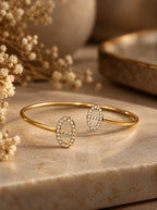 Athena Armreif in Gold mit ovalen Zirkonia-Enden auf heller Steinplatte neben Trockenblumen