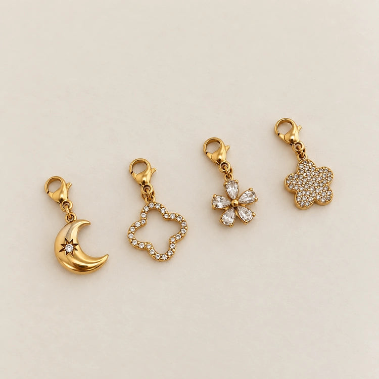 Kollektion aus vier 14k vergoldeten Edelstahl-Charms: Mond mit Stern-Gravur, Kleeblatt-Silhouette, kristallene Blütenform und voll besetzter Blüten-Anhänger.