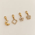 Kollektion aus vier 14k vergoldeten Edelstahl-Charms: Mond mit Stern-Gravur, Kleeblatt-Silhouette, kristallene Blütenform und voll besetzter Blüten-Anhänger.