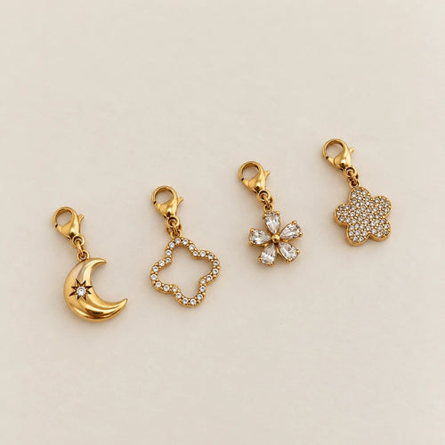 Kollektion aus vier 14k vergoldeten Edelstahl-Charms: Mond mit Stern-Gravur, Kleeblatt-Silhouette, kristallene Blütenform und voll besetzter Blüten-Anhänger.