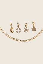 Vier vergoldete Charm-Anhänger mit Karabiner (Mond, Kleeblatt, Zirkonia-Blüte, Glitzerblume) arrangiert über einer groben Gliederkette.
