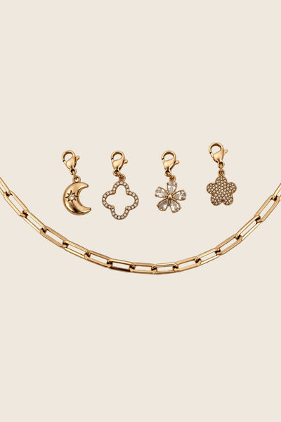 Vier vergoldete Charm-Anhänger mit Karabiner (Mond, Kleeblatt, Zirkonia-Blüte, Glitzerblume) arrangiert über einer groben Gliederkette.