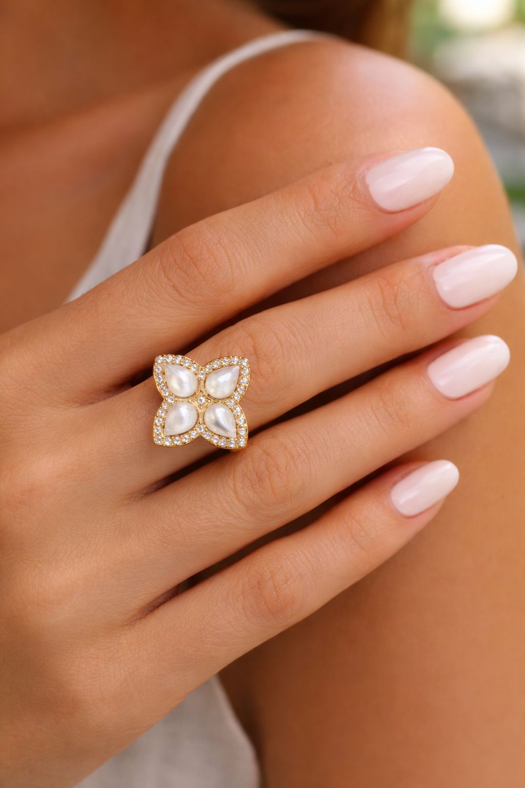 Nahaufnahme der Hand mit getragenem Statement Perlmutt-Ring 'Grande'. Zeigt das große Blumen-Motiv mit schimmernden Einlagen und Zirkonia-Halo kombiniert mit gepflegten Nude-Nägeln und einem weißen Oberteil.