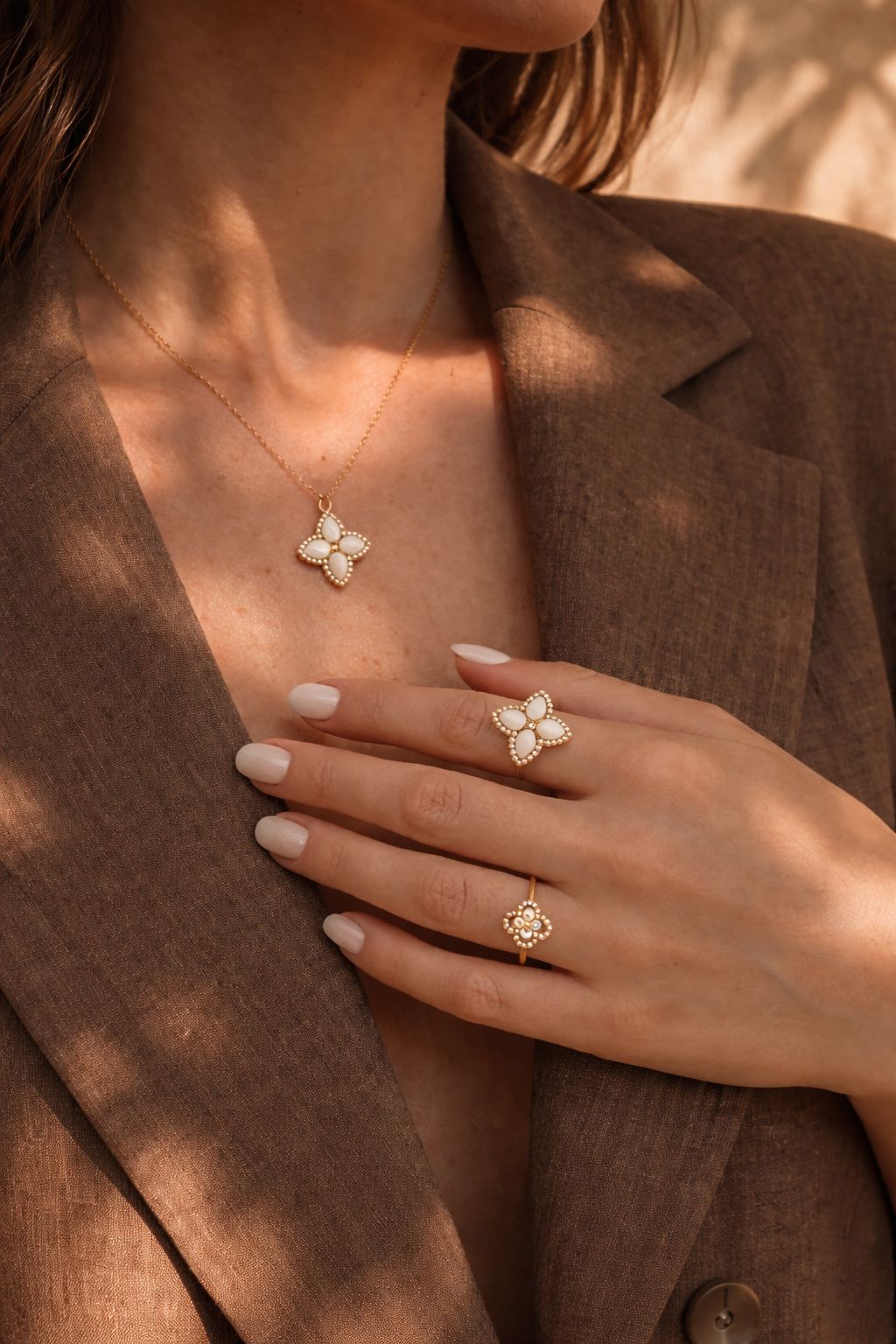 Cleo Ring am Finger getragen im Schmuck-Stacking. Die Aufnahme zeigt das Perlmutt-Kleeblatt-Design und die offene, verstellbare Ringschiene aus Gold, kombiniert mit der passenden Cleo Halskette und einem braunen Blazer.