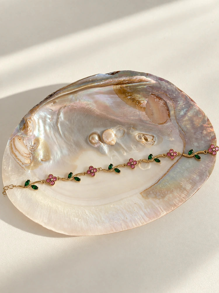 Gardenia Armband aus 14k vergoldetem Edelstahl mit rosa Blüten und grünen Blättern, drapiert auf einer großen, irisierenden Abalone-Muschel[cite: 5, 7]