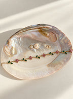 Gardenia Armband aus 14k vergoldetem Edelstahl mit rosa Blüten und grünen Blättern, drapiert auf einer großen, irisierenden Abalone-Muschel[cite: 5, 7]