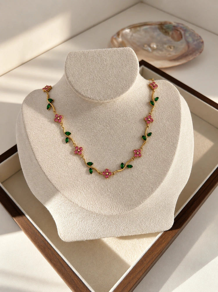 Gardenia Halskette aus 14k vergoldetem Edelstahl mit rosa Blüten und grünen Blättern präsentiert auf einer beigen Schmuckbüste
