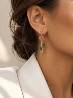 Hängende Ohrstecker im floralen Ranken-Design, gefertigt aus 14k vergoldetem Edelstahl. Ein geschwungener Stängel mit zwei grünen Navette-Blättern hängt unter einer kleinen Blüte aus rosa Zirkonia-Steinen. Getragen am Ohr einer Person mit heller Haut und dunklem, hochgestecktem Haar, die einen weißen Blazer trägt.