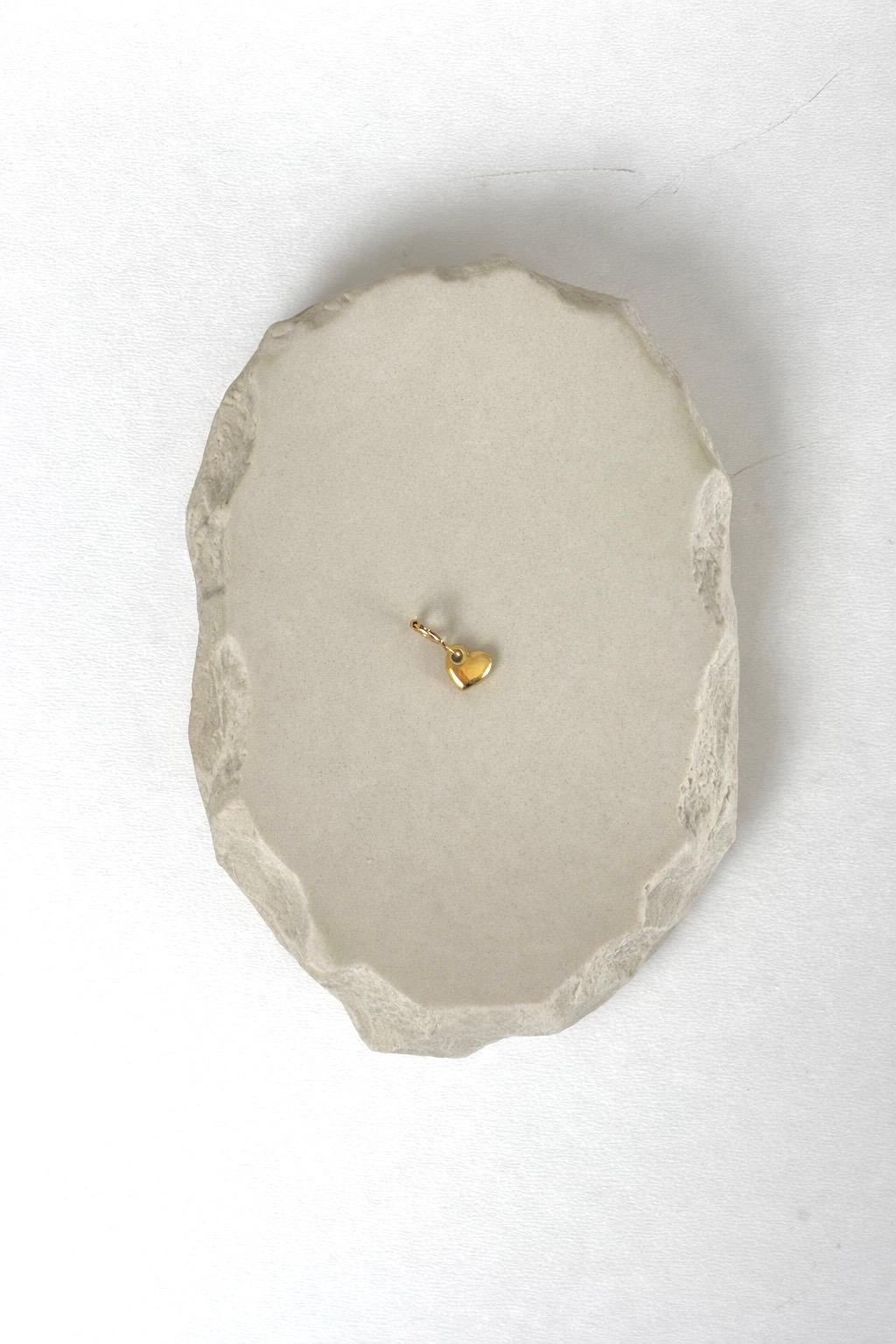 Minimalistischer goldener Herz-Anhänger mit Federring-Verschluss, präsentiert auf einem ovalen, hellgrauen Stein mit rauer Kante vor weißem Hintergrund.