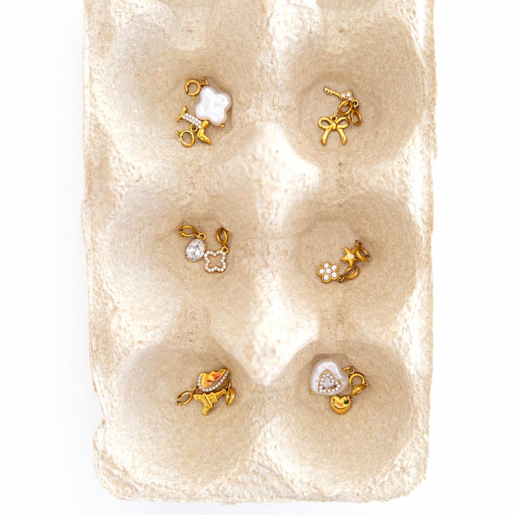 Vielfältige Auswahl an 14k vergoldeten Edelstahl-Charms, darunter Kleeblatt, Eiffelturm, Herz und Stern, arrangiert in einer minimalistischen, hellen Schmuckschale.