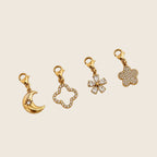 Moon Charm Halbmond-Anhänger neben weiteren Motiv-Charms aus vergoldetem Edelstahl auf hellem Untergrund.