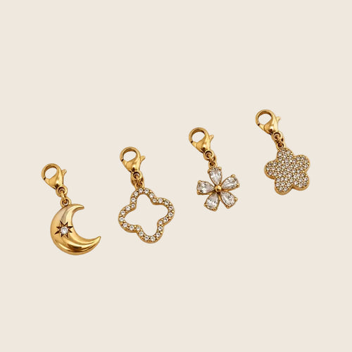 Moon Charm Halbmond-Anhänger neben weiteren Motiv-Charms aus vergoldetem Edelstahl auf hellem Untergrund.