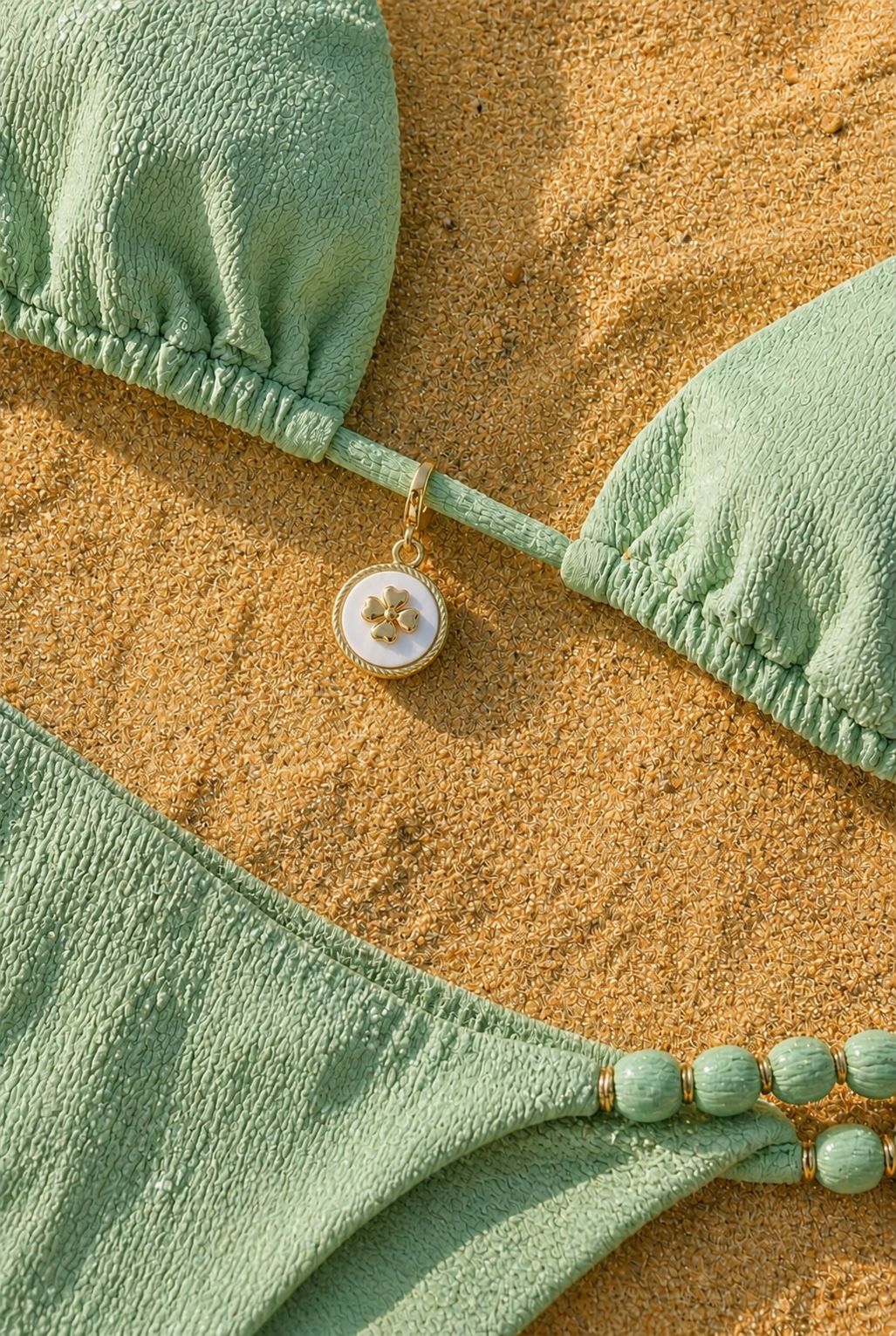 Goldener Ocean Luck Glücksbringer-Anhänger befestigt an einem hellgrünen Bikini im Sand.