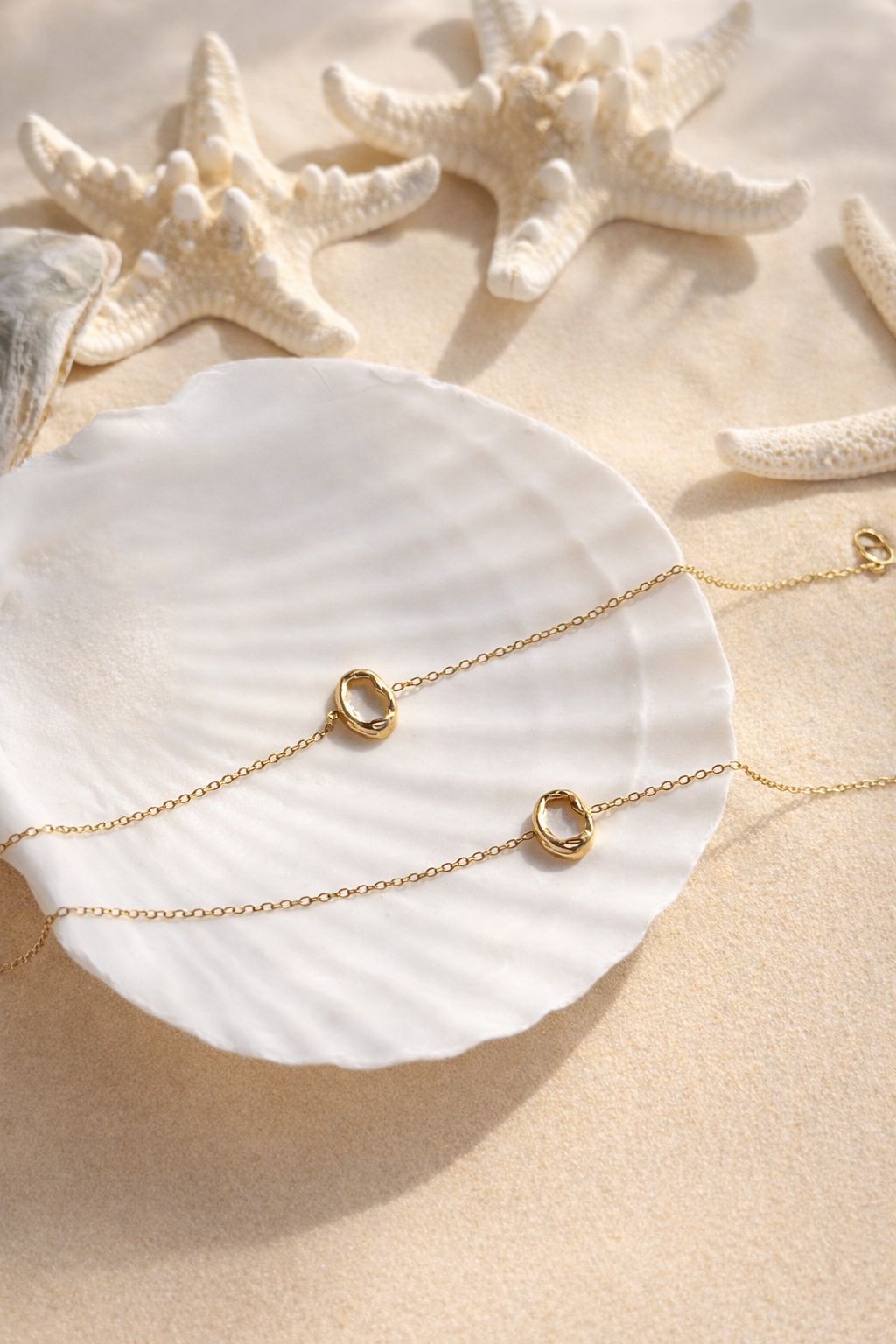 Zwei filigrane Aurora Armbänder aus vergoldetem Edelstahl, drapiert auf einer großen weißen Muschel im hellen Sand, flankiert von Seesternen zur Veranschaulichung des wasserfesten Designs.