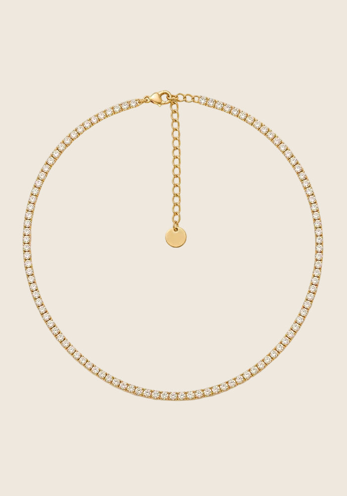 Shine Collier aus Edelstahl mit funkelnden Zirkonia-Steinen in Goldoptik auf beigem Hintergrund.