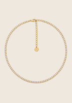 Shine Collier aus Edelstahl mit funkelnden Zirkonia-Steinen in Goldoptik auf beigem Hintergrund.